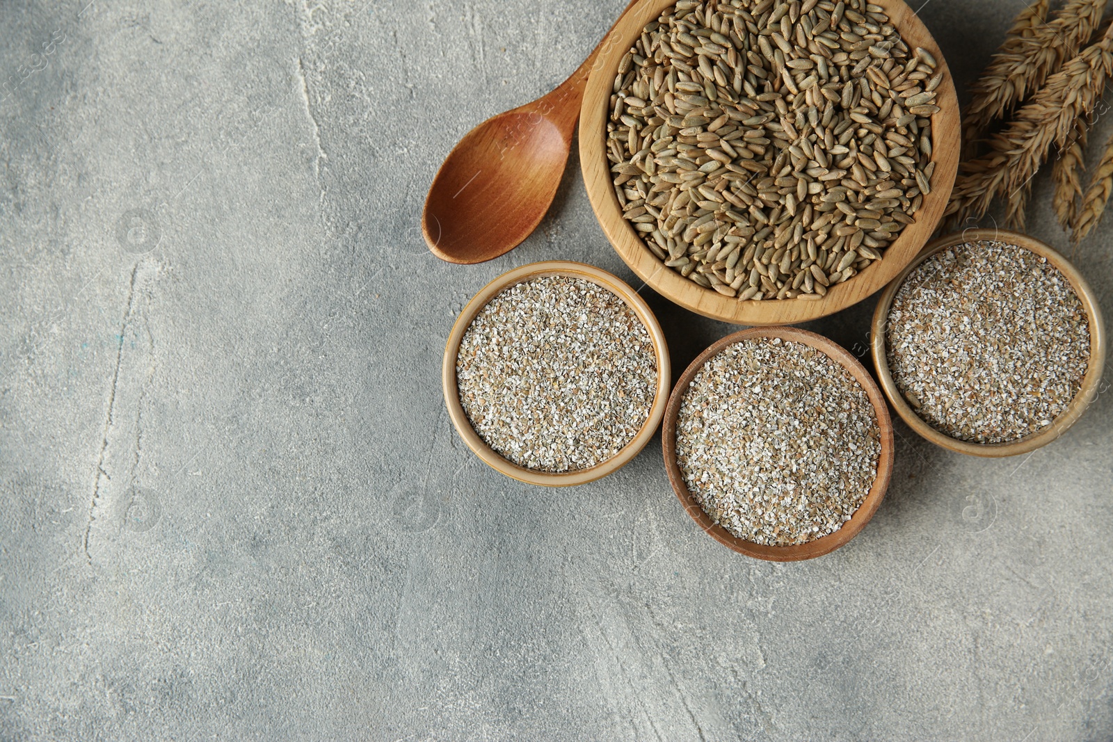 Photo of Fresh rye bran, kernels, spoon and spikelets on grey textured table, flat lay. Space for text