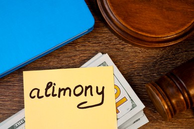 Photo of Card with word Alimony, dollar banknotes, gavel and notebook on wooden table, top view