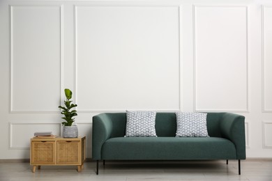 Photo of Stylish sofa with cushions, plant and books on side table near white wall