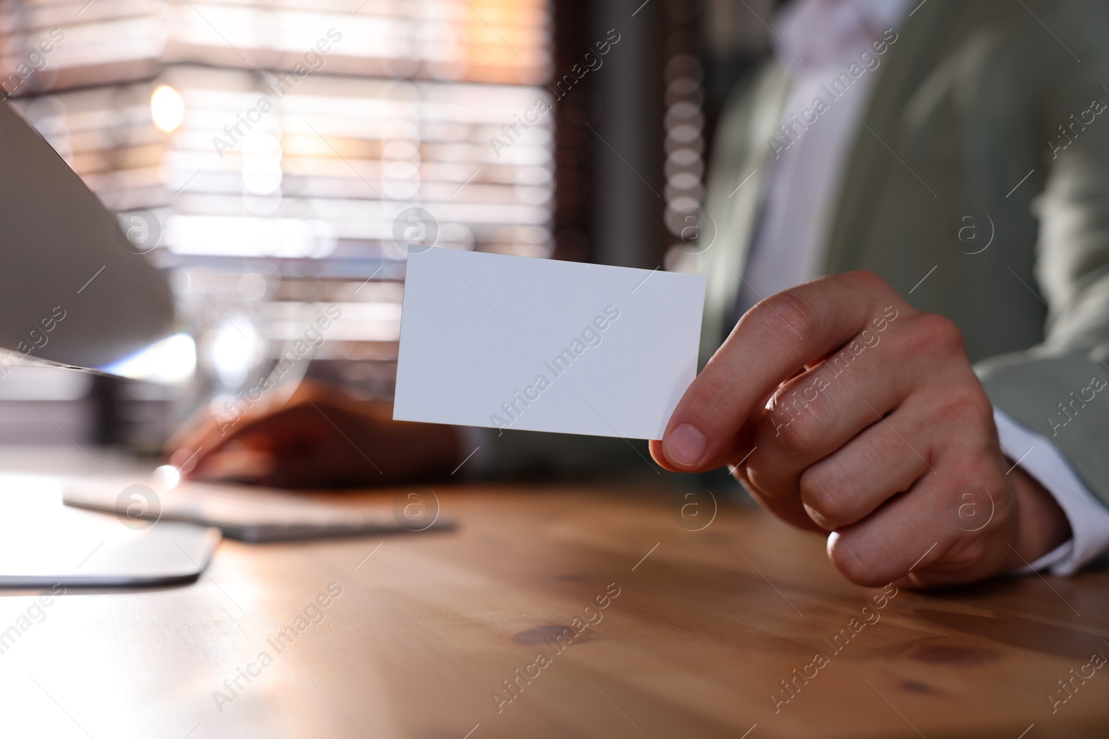 Photo of Man holding blank business card while working with computer at table in office, closeup. Mockup for design