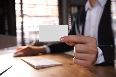 Man holding blank business card while working with computer at table in office, closeup. Mockup for design