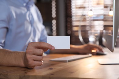 Photo of Man holding blank business card while working with computer at table in office, closeup. Mockup for design