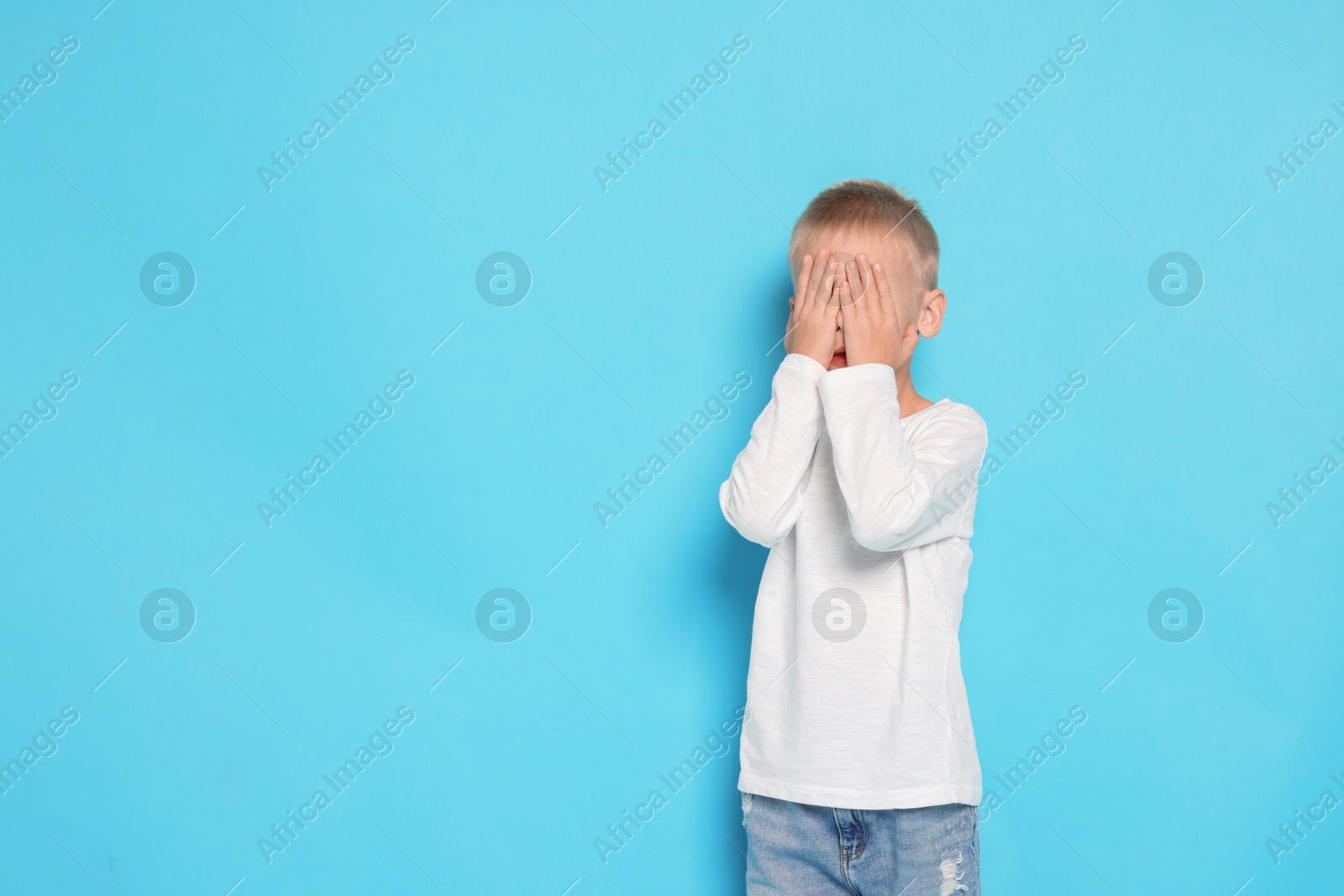 Photo of Scared little boy covering face with hands on light blue background. Space for text