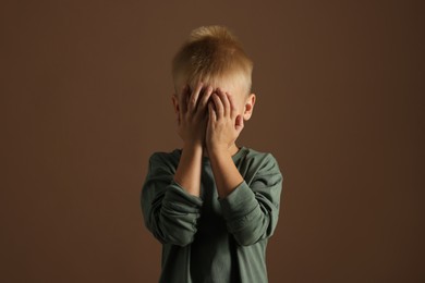 Scared little boy covering face with hands on brown background