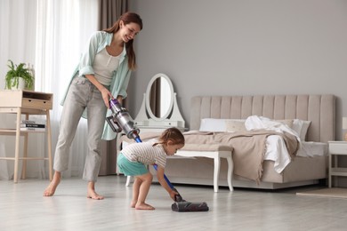 Photo of Smiling young woman and her daughter cleaning floor with cordless vacuum cleaner in bedroom