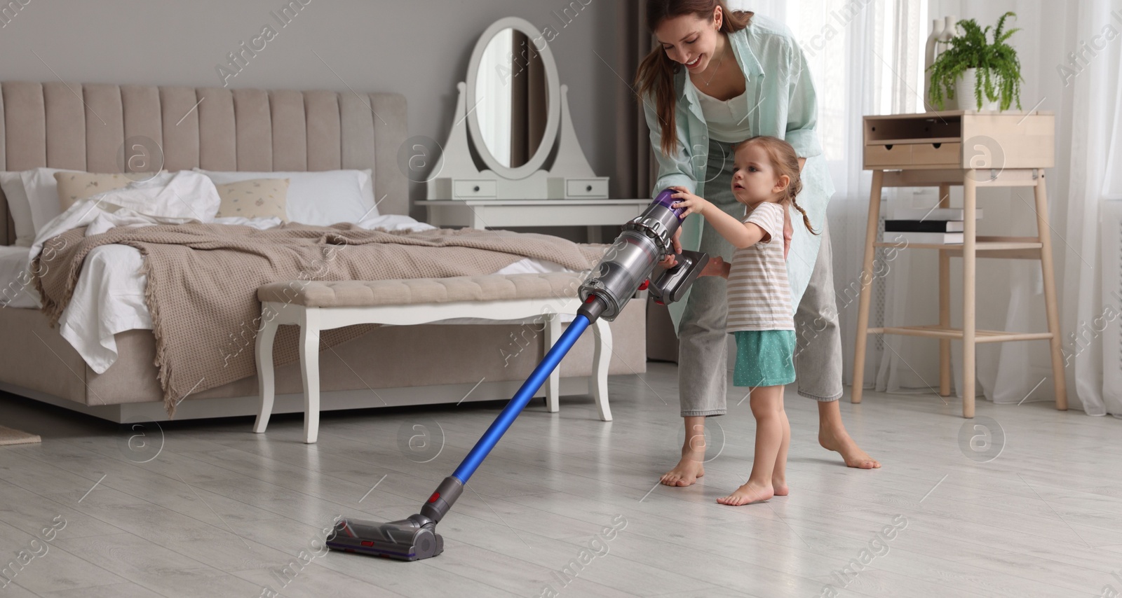 Photo of Smiling young woman and her daughter cleaning floor with cordless vacuum cleaner in bedroom