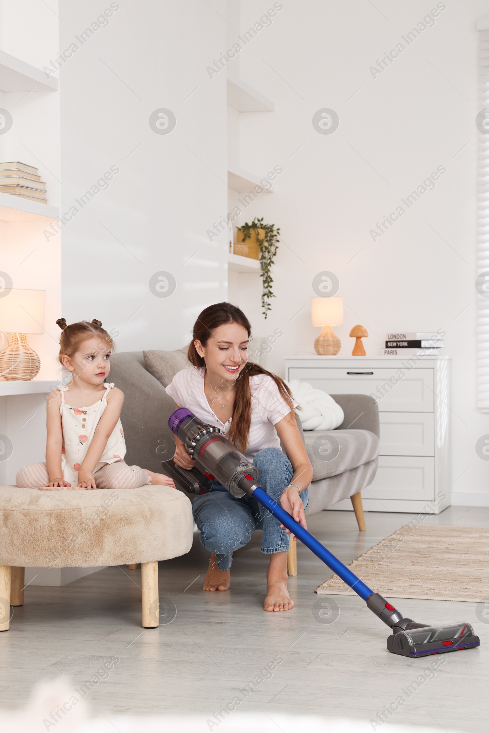 Photo of Smiling young woman and her daughter cleaning floor with cordless vacuum cleaner at home