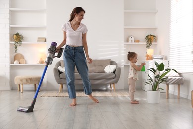 Smiling young woman cleaning floor with cordless vacuum cleaner while her daughter spraying houseplant at home