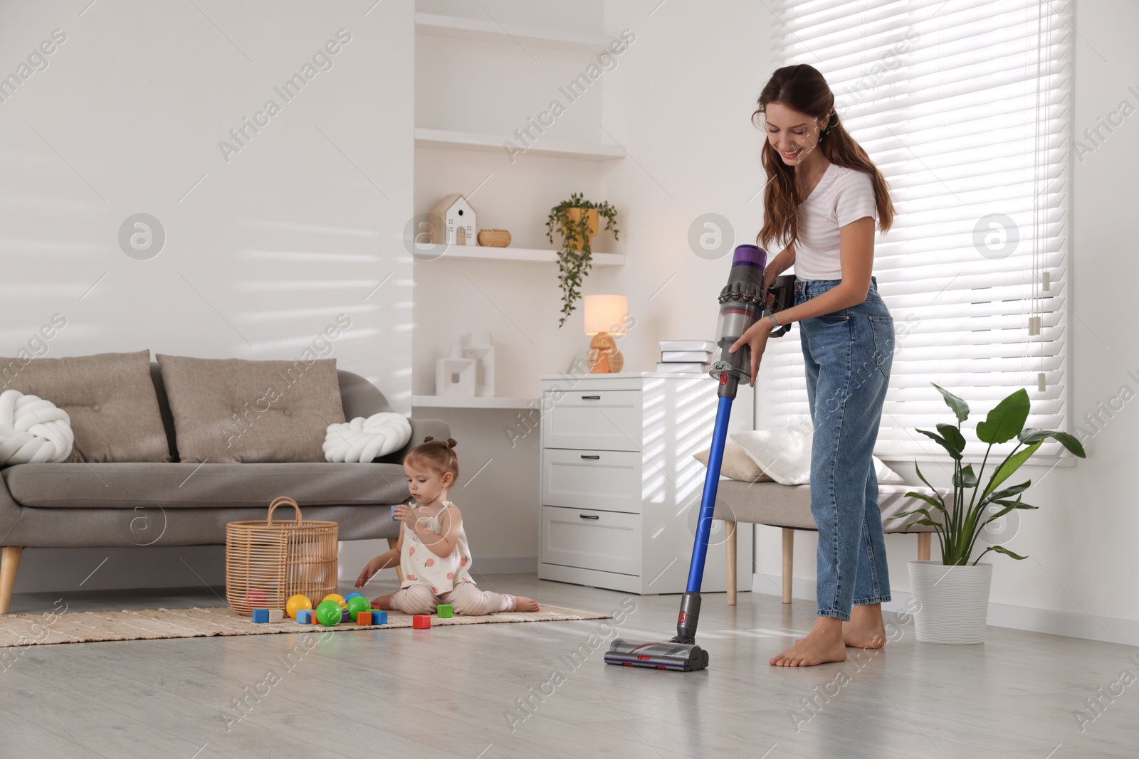 Photo of Smiling young woman cleaning floor with cordless vacuum cleaner while her daughter playing with toys at home