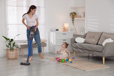 Smiling young woman cleaning floor with cordless vacuum cleaner while her daughter playing with toys at home