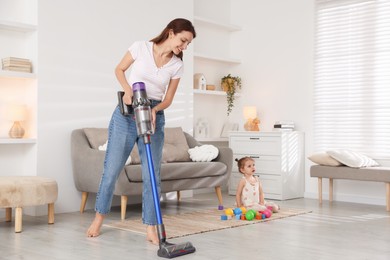 Smiling young woman cleaning floor with cordless vacuum cleaner while her daughter playing with toys at home