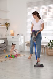 Photo of Smiling young woman cleaning floor with cordless vacuum cleaner while her daughter playing with toys at home