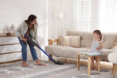 Young woman cleaning rug with cordless vacuum cleaner while her daughter drawing at home