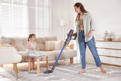 Smiling young woman cleaning rug with cordless vacuum cleaner while her daughter drawing at home