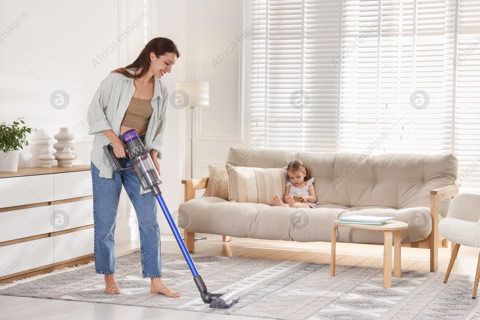 Photo of Smiling young woman cleaning rug with cordless vacuum cleaner while her daughter sitting on sofa at home