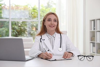 Photo of Doctor with stethoscope, laptop and clipboard at table in clinic