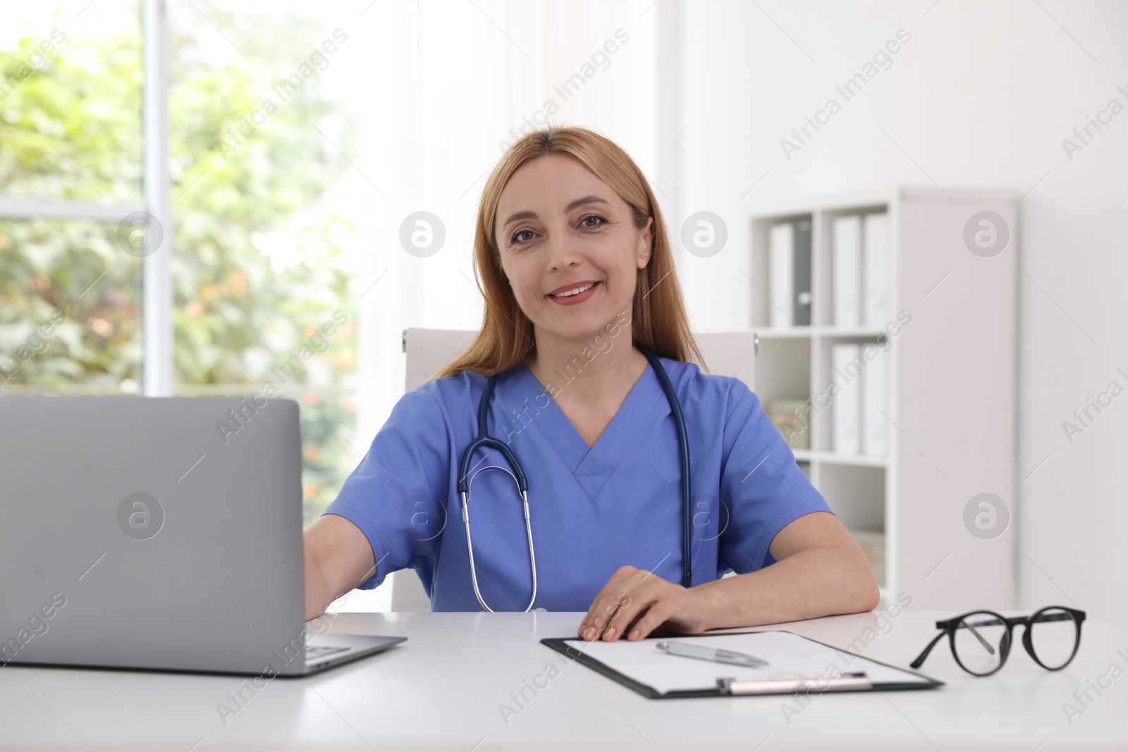 Photo of Doctor with stethoscope, laptop and clipboard at table in clinic