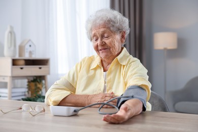 Senior woman measuring blood pressure at wooden table indoors