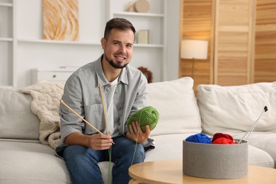 Man with green yarn and knitting needles on sofa at home