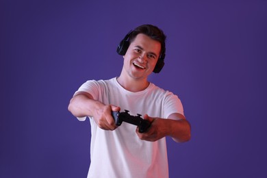 Happy young man in headphones playing video games with controller on violet background