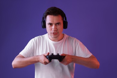 Young man in headphones playing video games with controller on violet background