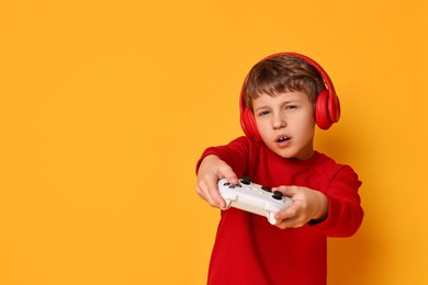 Cute little boy in headphones playing video game with controller on orange background. Space for text