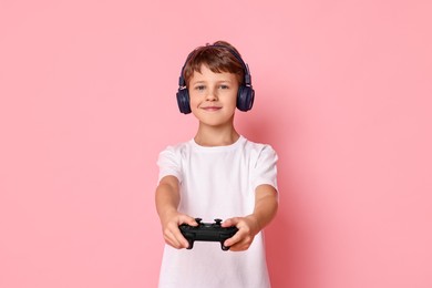 Cute little boy in headphones playing video game with controller on pink background