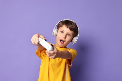 Cute little boy in headphones playing video game with controller on purple background