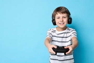 Happy little boy in headphones playing video game with controller on light blue background. Space for text