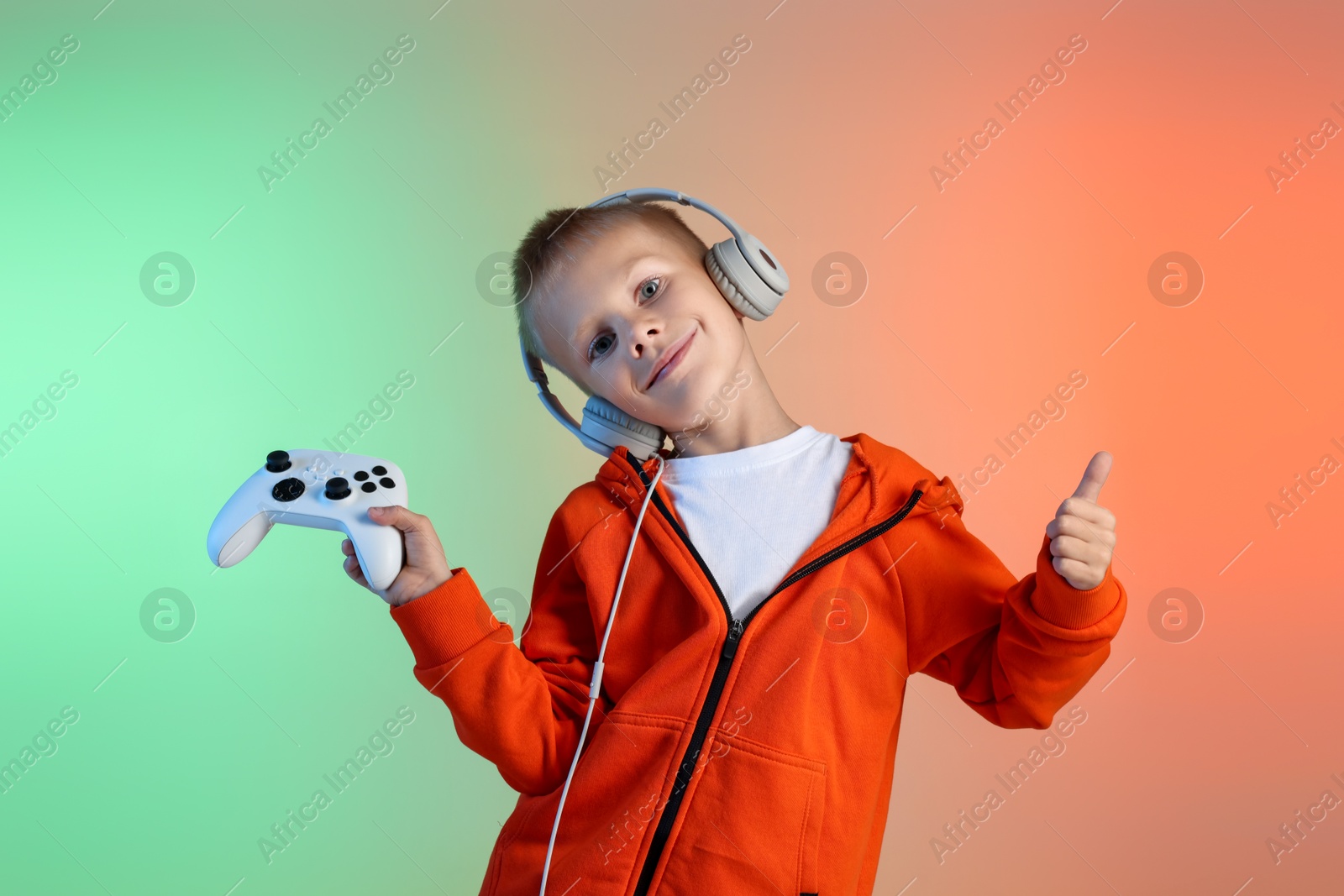 Photo of Cute little boy in headphones with controller showing thumbs up on color background