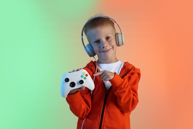 Photo of Cute little boy in headphones pointing at controller on color background