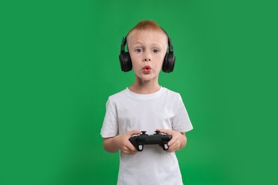 Cute little boy in headphones playing video game with controller on green background