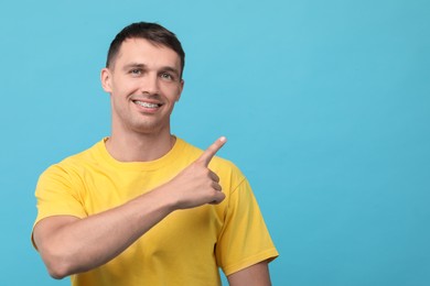 Smiling man with dental braces pointing at something on light blue background. Space for text