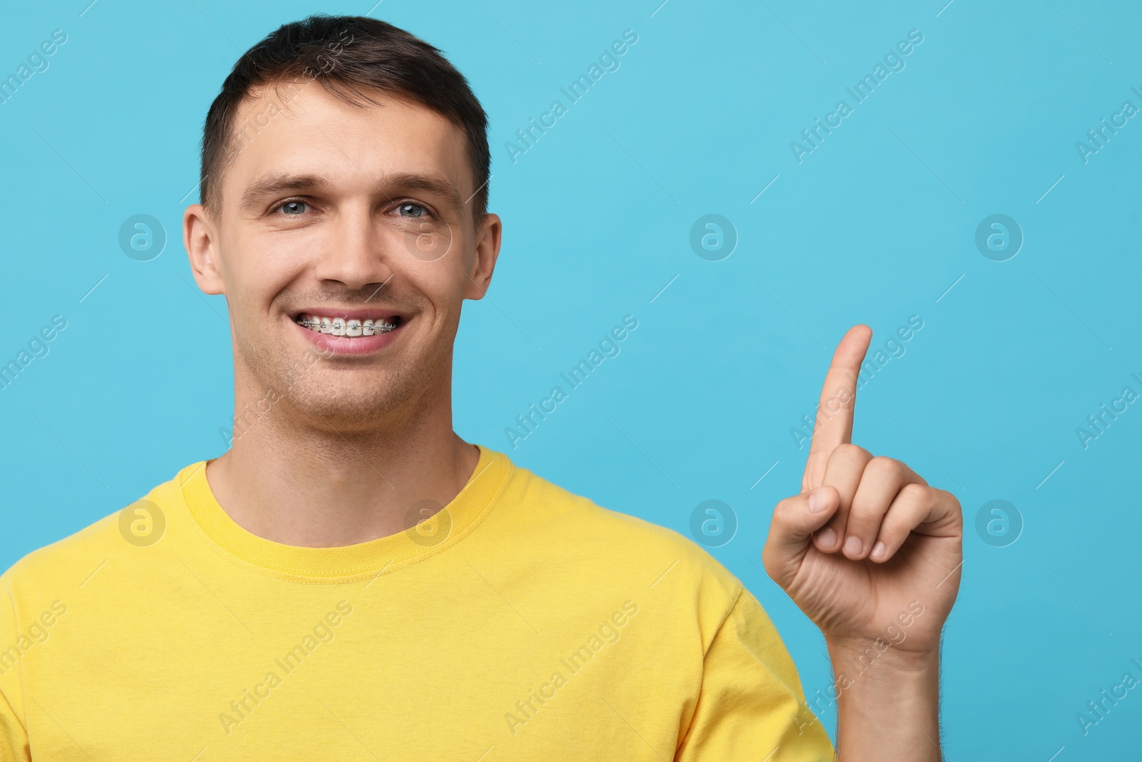 Photo of Smiling man with dental braces pointing at something on light blue background. Space for text