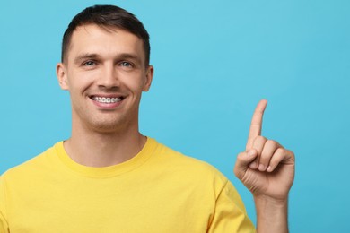 Smiling man with dental braces pointing at something on light blue background. Space for text