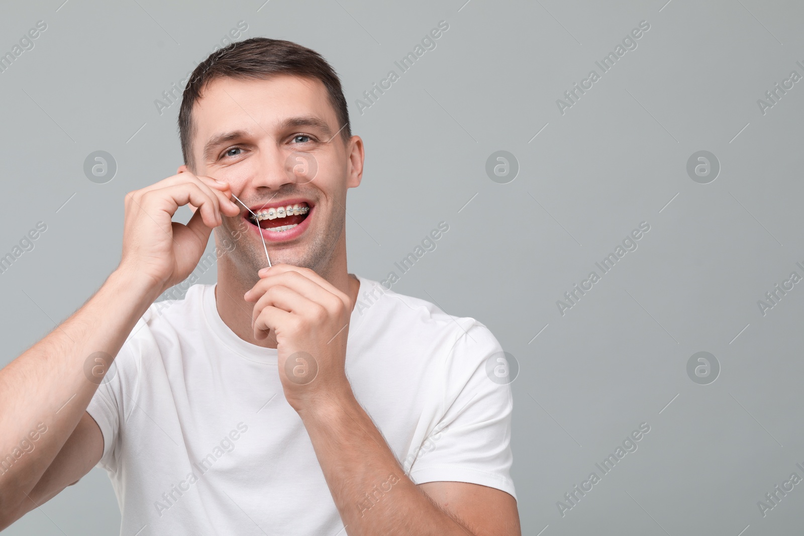 Photo of Happy man with braces cleaning teeth using dental floss on grey background. Space for text