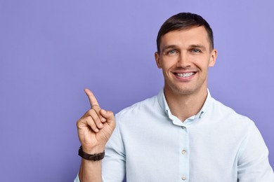 Photo of Smiling man with dental braces pointing at something on violet background. Space for text