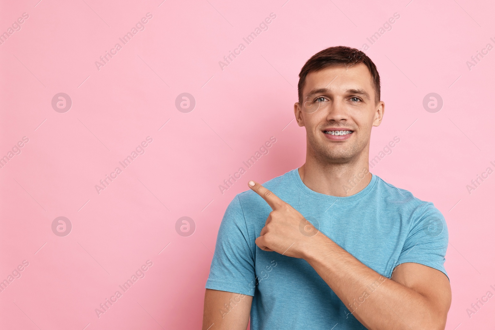 Photo of Smiling man with dental braces pointing at something on pink background. Space for text