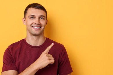 Smiling man with dental braces pointing at something on yellow background. Space for text