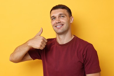 Smiling man with dental braces showing thumbs up on yellow background