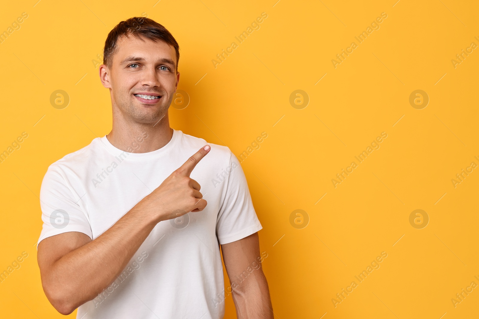 Photo of Smiling man with dental braces pointing at something on yellow background. Space for text