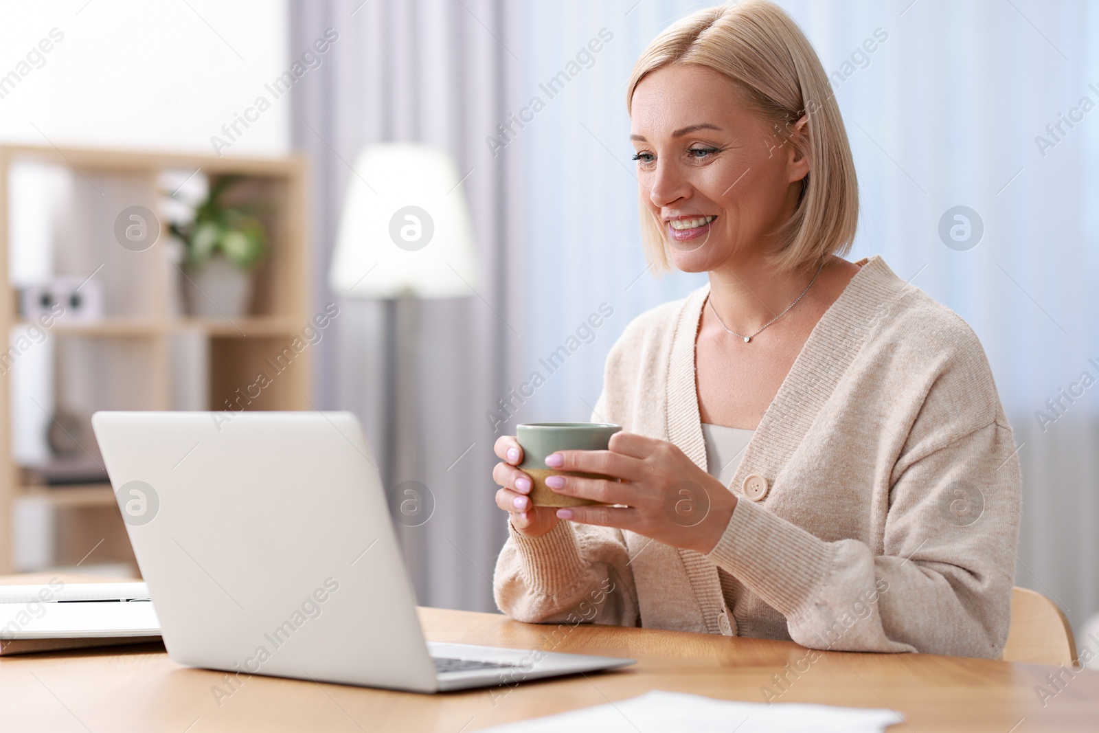 Photo of Happy middle aged woman with cup of drink using laptop at table indoors