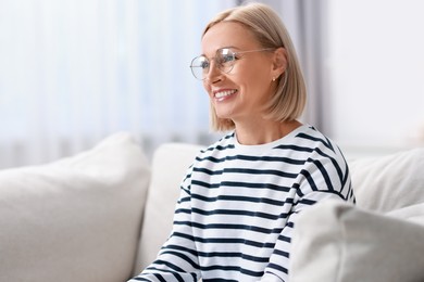 Happy middle aged woman in glasses on sofa indoors. Space for text