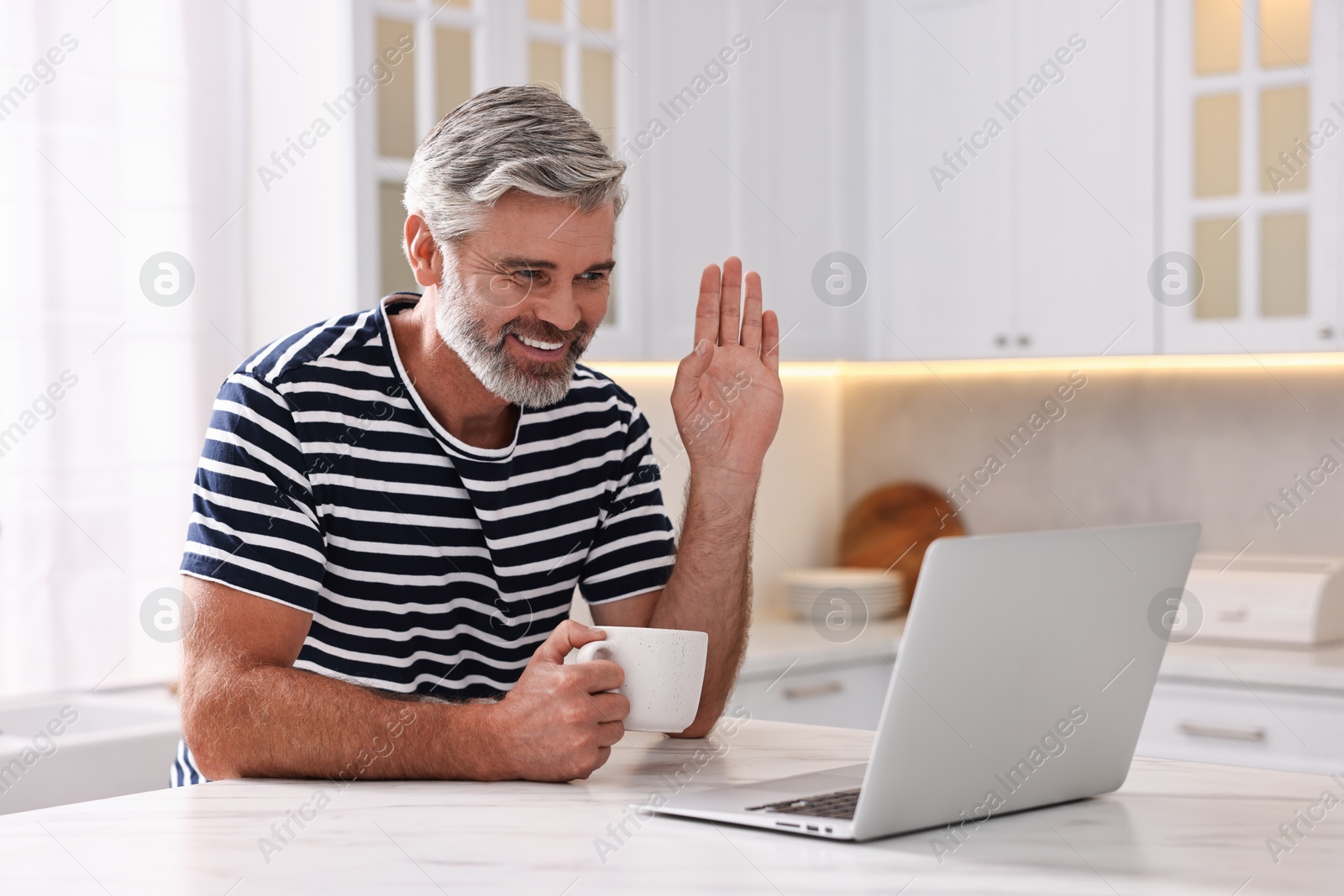 Photo of Happy middle aged man with cup of drink having video chat via laptop at white marble table in kitchen