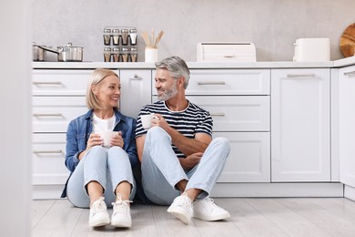 Happy middle aged couple with cups of drink on floor in kitchen, space for text