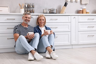 Happy middle aged couple with cups of drink on floor in kitchen, space for text