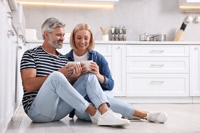 Photo of Happy middle aged couple with cups of drink on floor in kitchen, space for text