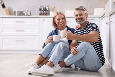 Photo of Happy middle aged couple with cups of drink on floor in kitchen, space for text