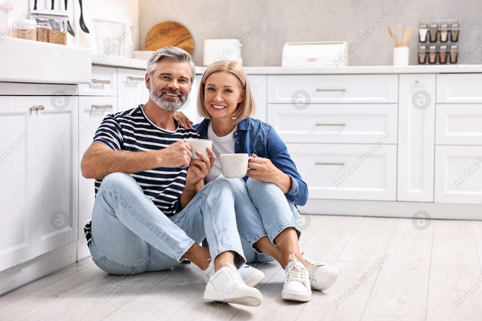 Photo of Happy middle aged couple with cups of drink on floor in kitchen, space for text