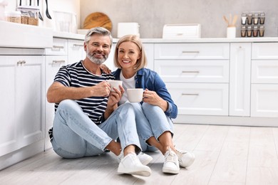 Happy middle aged couple with cups of drink on floor in kitchen, space for text